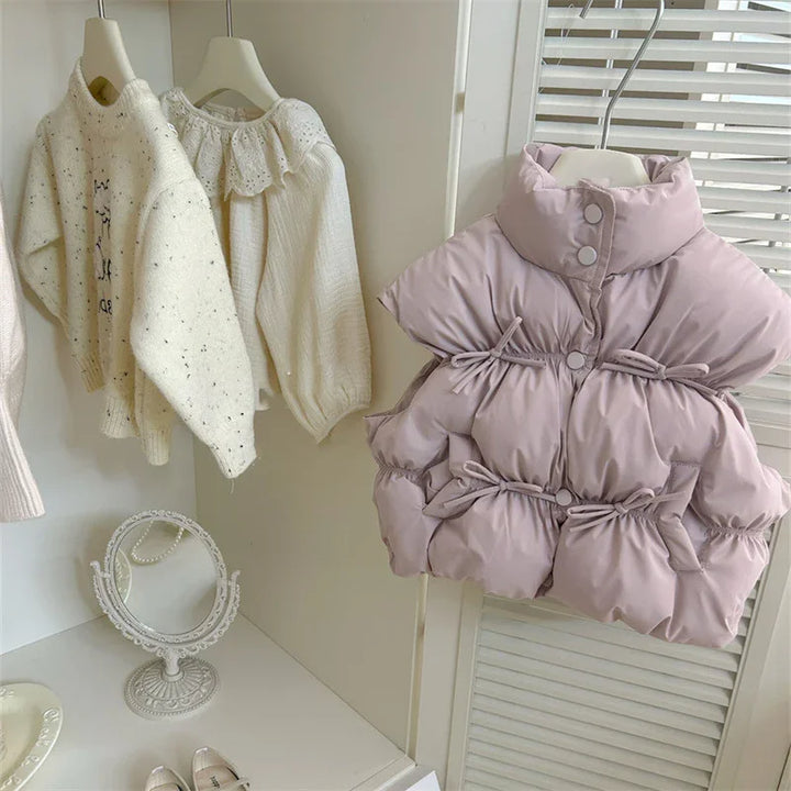Light purple padded sleeveless jacket with buttons and drawstrings hanging on a rack beside cream knit sweaters