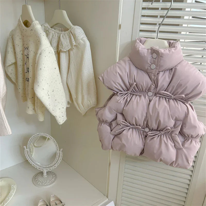 Lavender padded sleeveless jacket with buttons and ties hanging next to white knitted sweaters and lace blouse