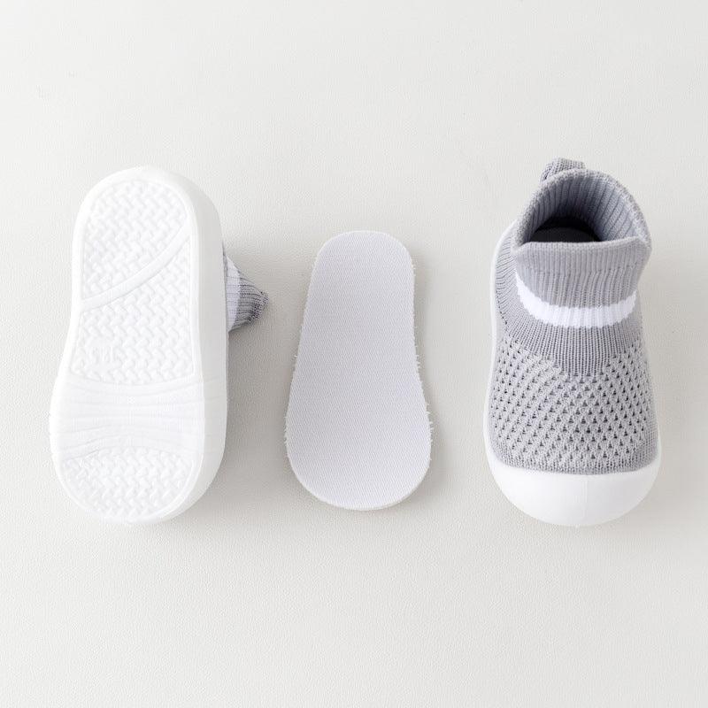 Gray knit baby sock sneakers with white rubber soles and a removable insole on a light background