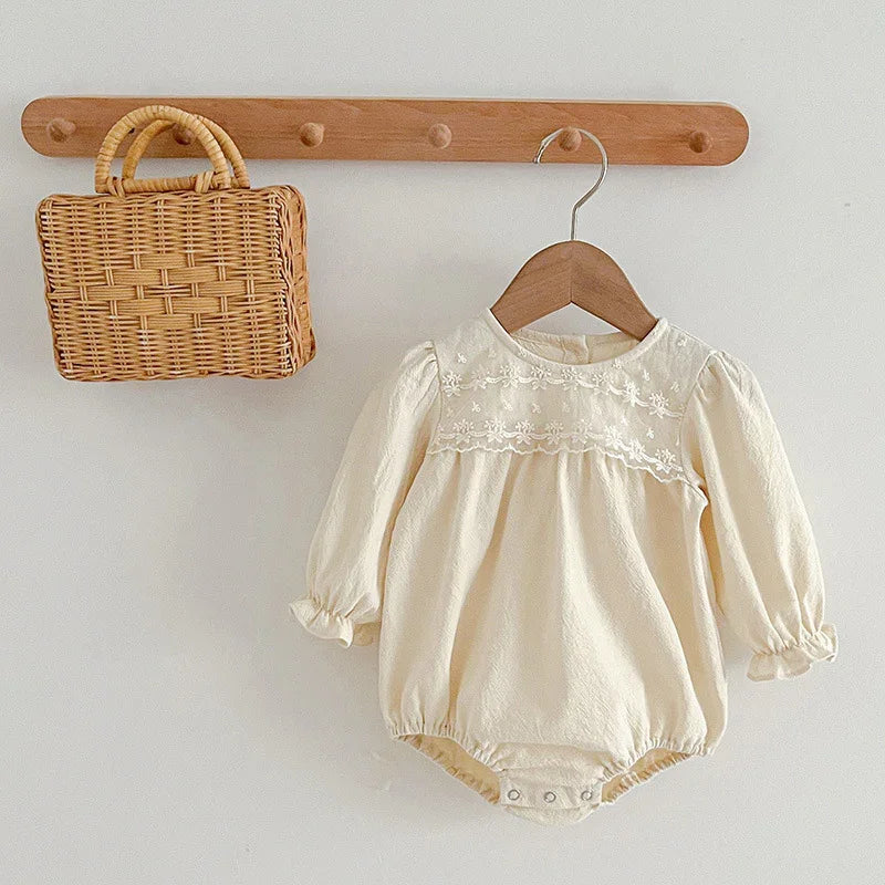 Cream baby romper with floral embroidery hanging on wooden hanger next to wicker handbag on wall hook