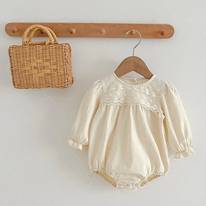 Cream baby romper with floral embroidery hanging on wooden hanger next to wicker handbag on wall hook