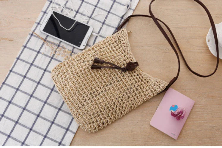 Handwoven straw crossbody bag with brown strap on wooden table with phone, pink power bank, and checkered cloth