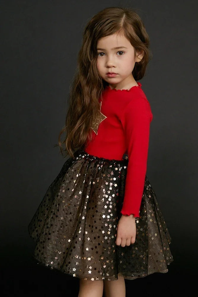 Young girl in red long-sleeve top with gold star detail and black star-patterned tulle skirt on dark background