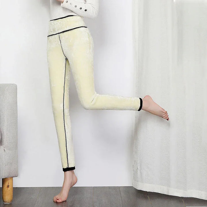 woman wearing light yellow fleece leggings with black trim, standing on one foot indoors by curtain and gray chair
