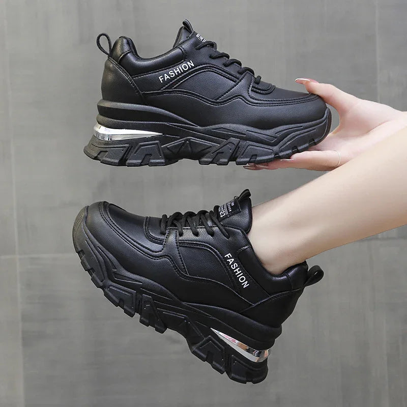 Pair of black chunky platform sneakers with FASHION branding held against gray tiled background