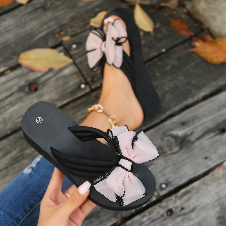 Black slip-on sandals with large pink bows worn on feet over wooden deck with autumn leaves