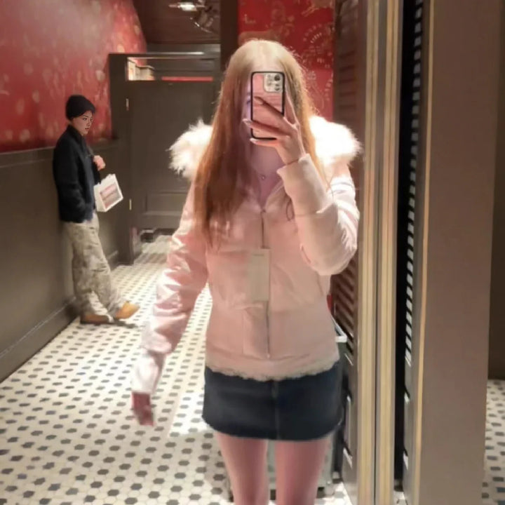 Mirror selfie of woman in pink fur-collared jacket and denim skirt in a stylish restroom with red wallpaper