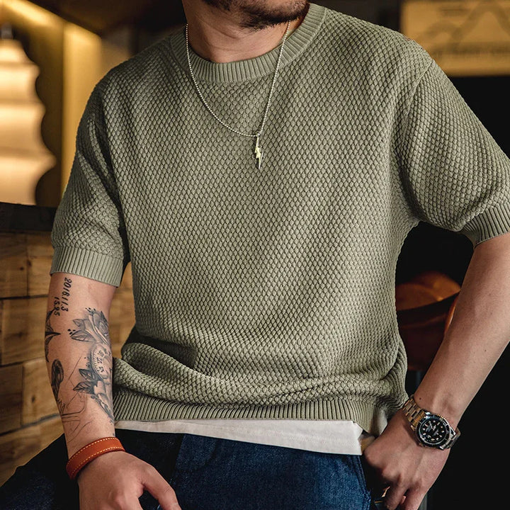 Man wearing textured olive green short-sleeve sweater with silver lightning pendant necklace, tattoos, and wristwatch