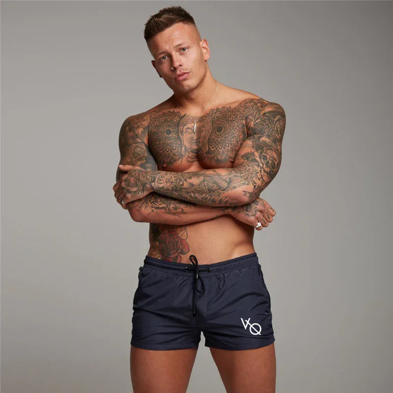 muscular tattooed man wearing navy VQ branded shorts posing with crossed arms on gray background
