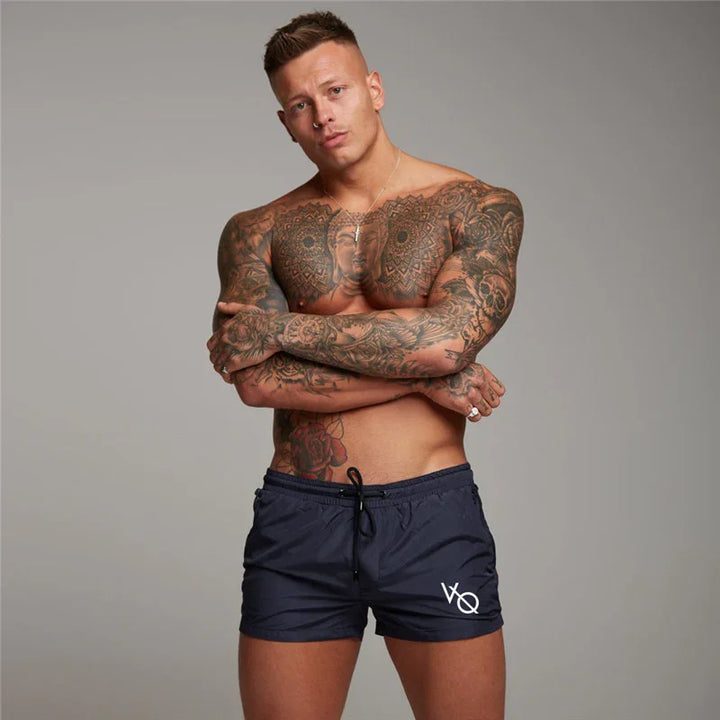 muscular tattooed man wearing navy VQ branded shorts posing with crossed arms on gray background