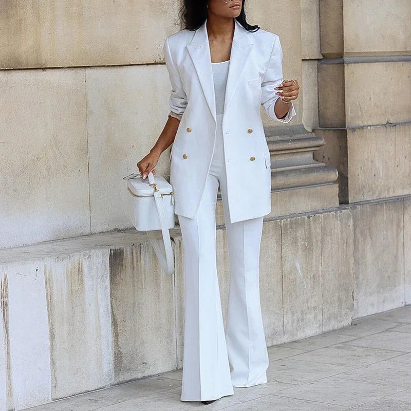Woman wearing stylish white double-breasted blazer and flared pants, holding white handbag against stone wall