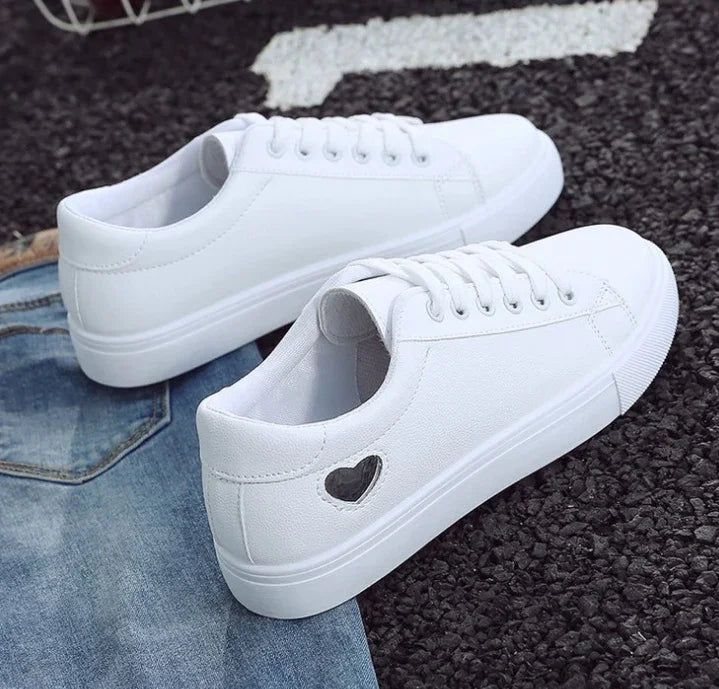 White casual sneakers with a small heart-shaped cutout detail on the side, placed on a denim jacket and black textured surface