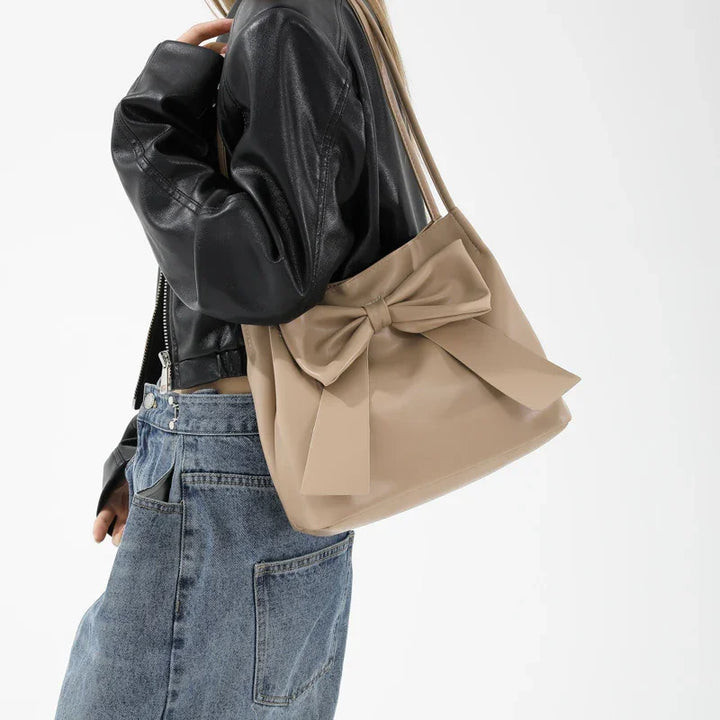 Woman wearing black leather jacket and blue jeans carrying beige tote bag with large bow detail