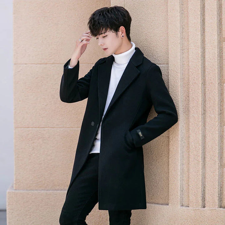 Young man wearing black long coat and white turtleneck sweater leaning against textured beige wall