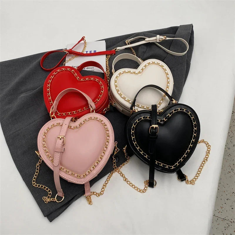 Heart-shaped leather handbags in red, pink, white, and black with gold chain accents on gray fabric