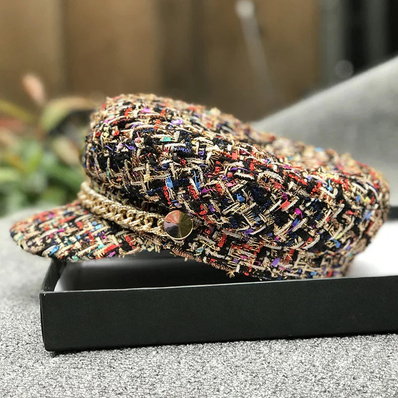 Colorful tweed newsboy cap with gold chain detail resting on black box on textured gray surface