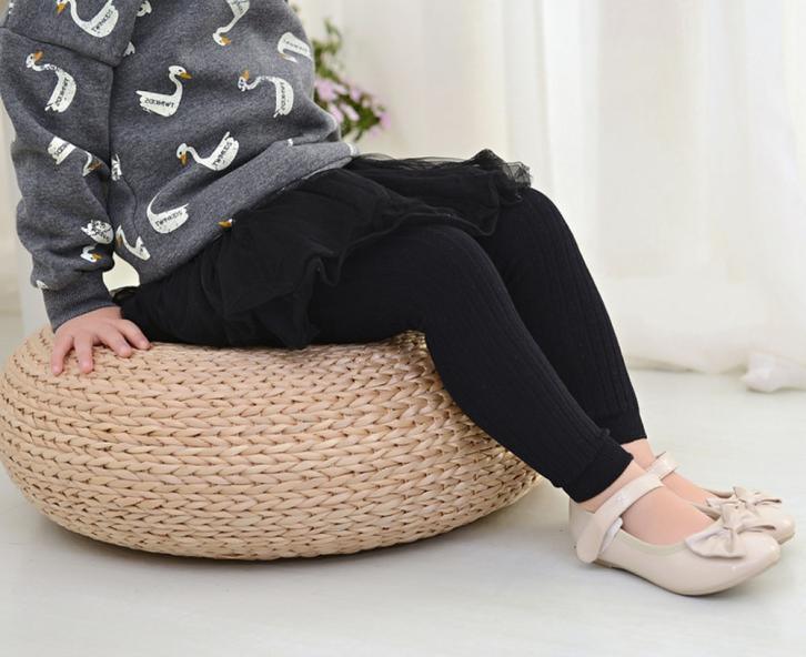 Child wearing grey swan-print sweater, black leggings, black skirt, and cream bow shoes sitting on woven round cushion