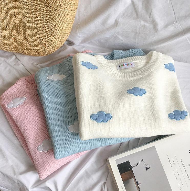 Stack of pastel sweaters with blue and white cloud embroidery on white fabric background
