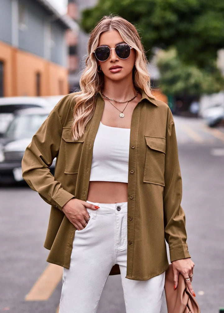 Woman wearing olive green button-up shirt, white crop top, white pants, sunglasses, holding clutch on urban street