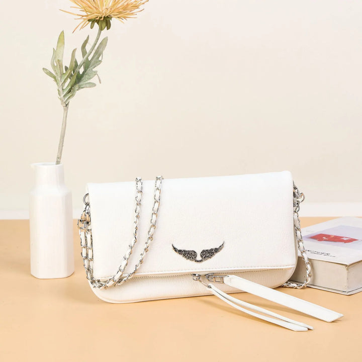White leather shoulder bag with silver chain strap and black wing emblem on beige table with flower vase and book