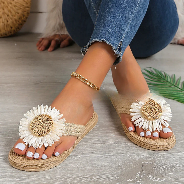 Close-up of feet wearing beige woven sandals with large white daisy flower decorations and gold anklet on wooden floor