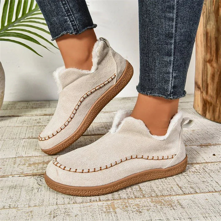 Beige fleece-lined moccasin slippers with stitched details worn with dark blue jeans on wooden floor