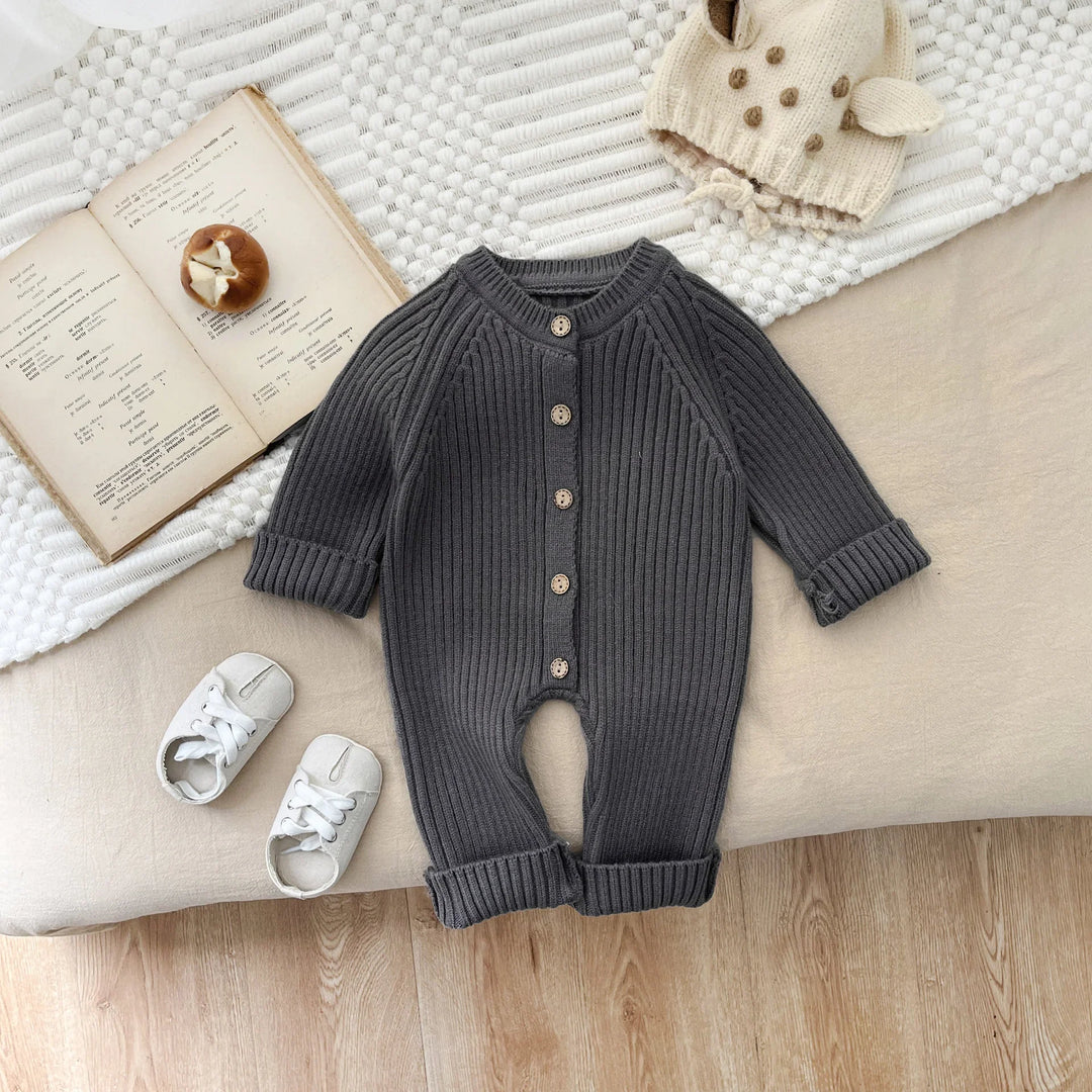 Gray ribbed baby onesie with wooden buttons, white baby shoes, knitted cream hat, and open book on beige and white textured surfaces