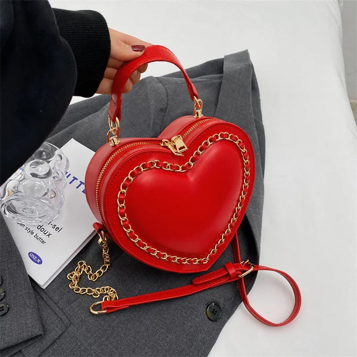 Red heart-shaped handbag with gold chain detail and zipper, held over gray coat and magazine