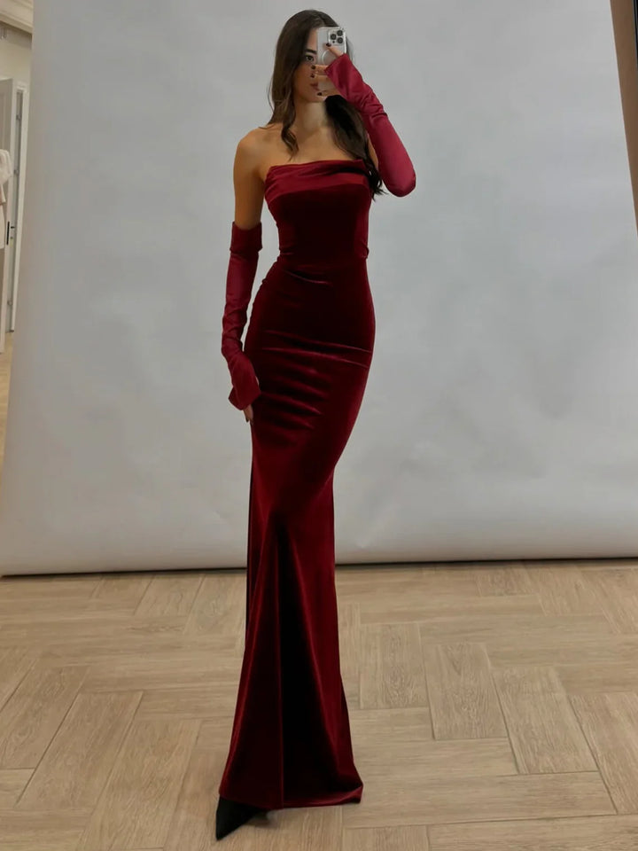 Woman in a fitted strapless burgundy velvet gown with matching long gloves, posing in front of a gray backdrop