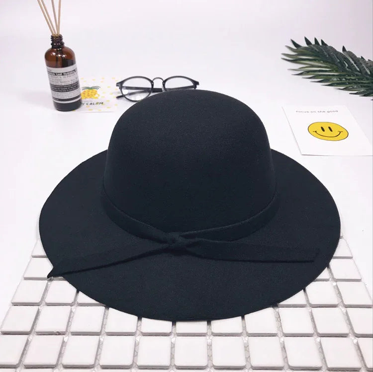 Black wide-brimmed felt hat with bow detail on white tiled surface, styled with glasses and decorative items