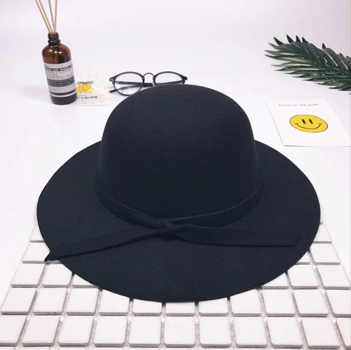 Black wide-brimmed felt hat with bow detail on white tiled surface, styled with glasses and decorative items