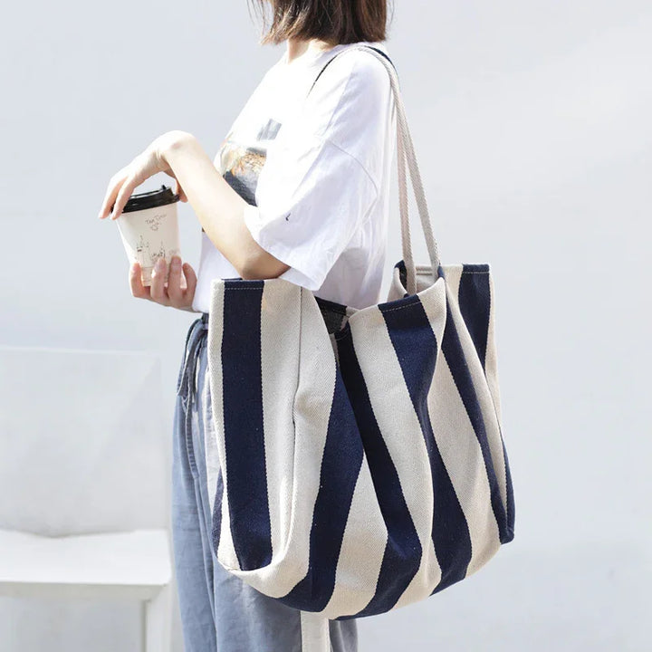Woman holding coffee cup wearing white t-shirt and blue-gray pants with navy blue and white striped tote bag