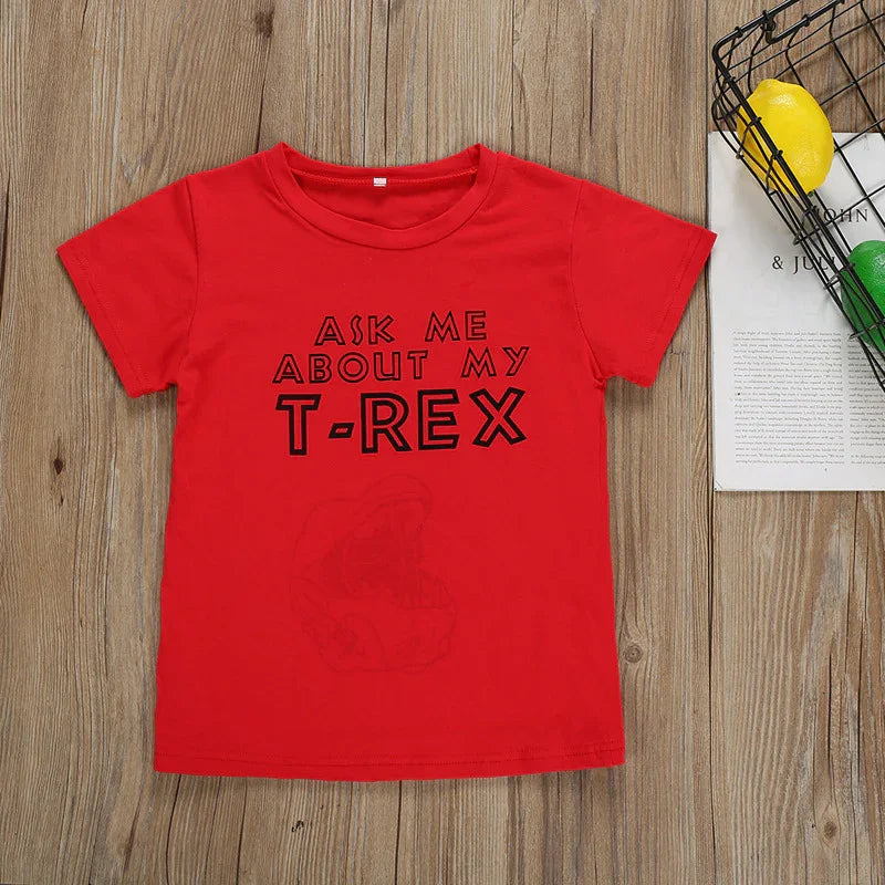 Red kids T-shirt with text Ask me about my T-Rex on wooden surface next to wire basket with lemons