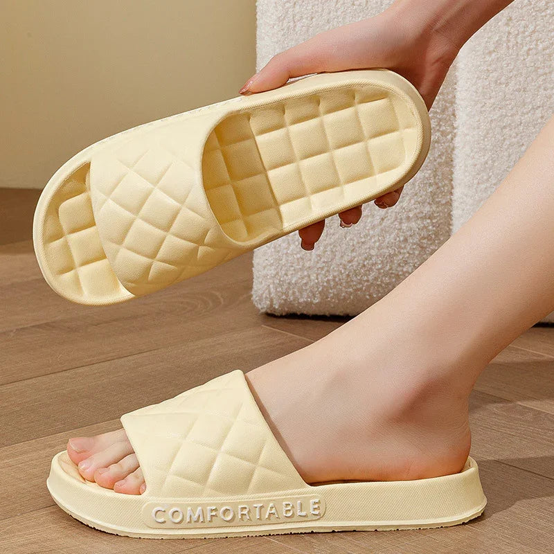 Close-up of beige quilted slide sandals with cushioned sole, one worn and one held by hand, on wooden floor