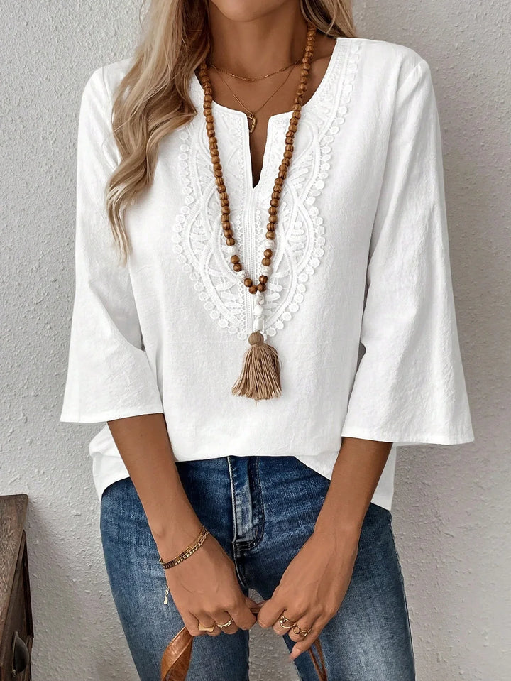 Woman wearing white embroidered blouse with 3/4 bell sleeves and blue jeans, accessorized with wooden beaded necklace and gold jewelry