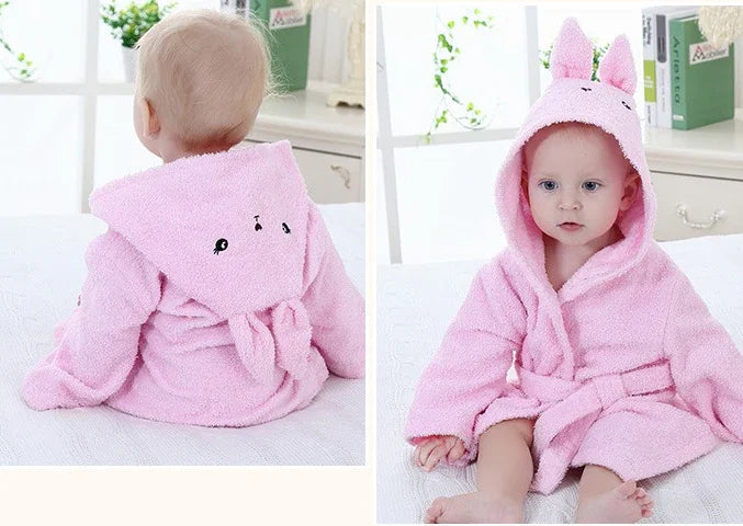 Baby wearing a pink hooded bathrobe with bunny ears and embroidered face, sitting on a white bed