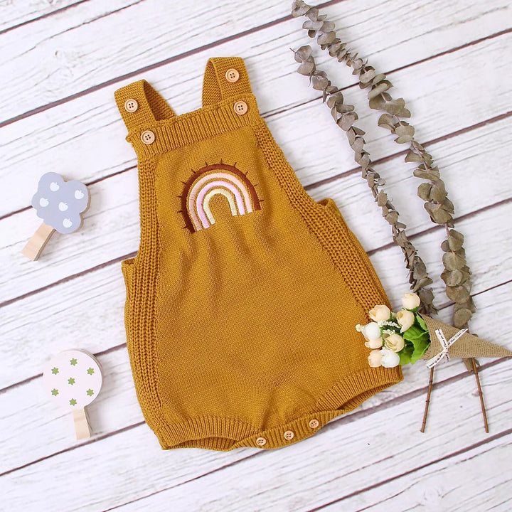 Mustard yellow knitted baby romper with rainbow embroidery on wooden background with decorative flowers