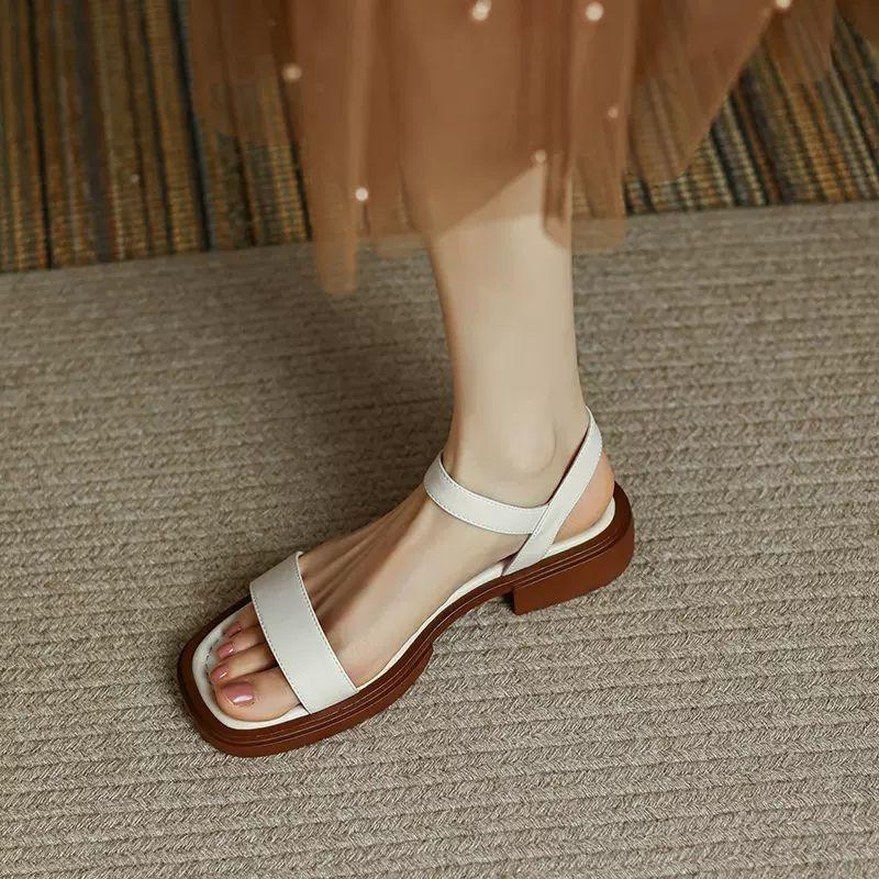 Woman wearing stylish white flat sandals with brown sole on carpeted floor