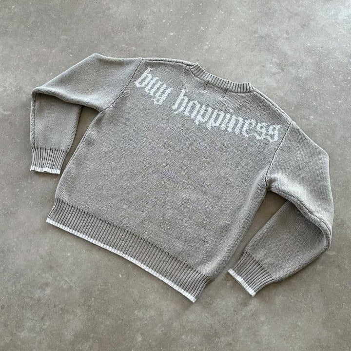 Gray knit sweater with white trim displaying 'buy happiness' text on the back, laid on concrete floor
