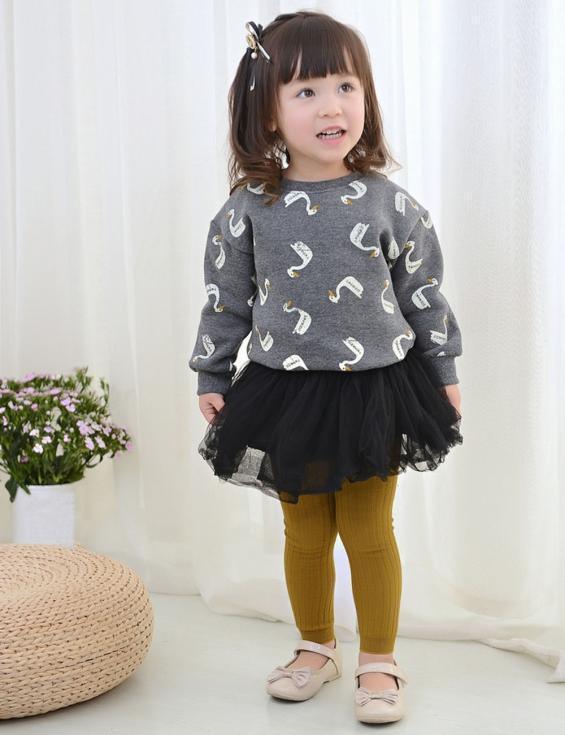 Toddler girl wearing grey swan print sweater, black tutu skirt, mustard leggings, and beige shoes indoors