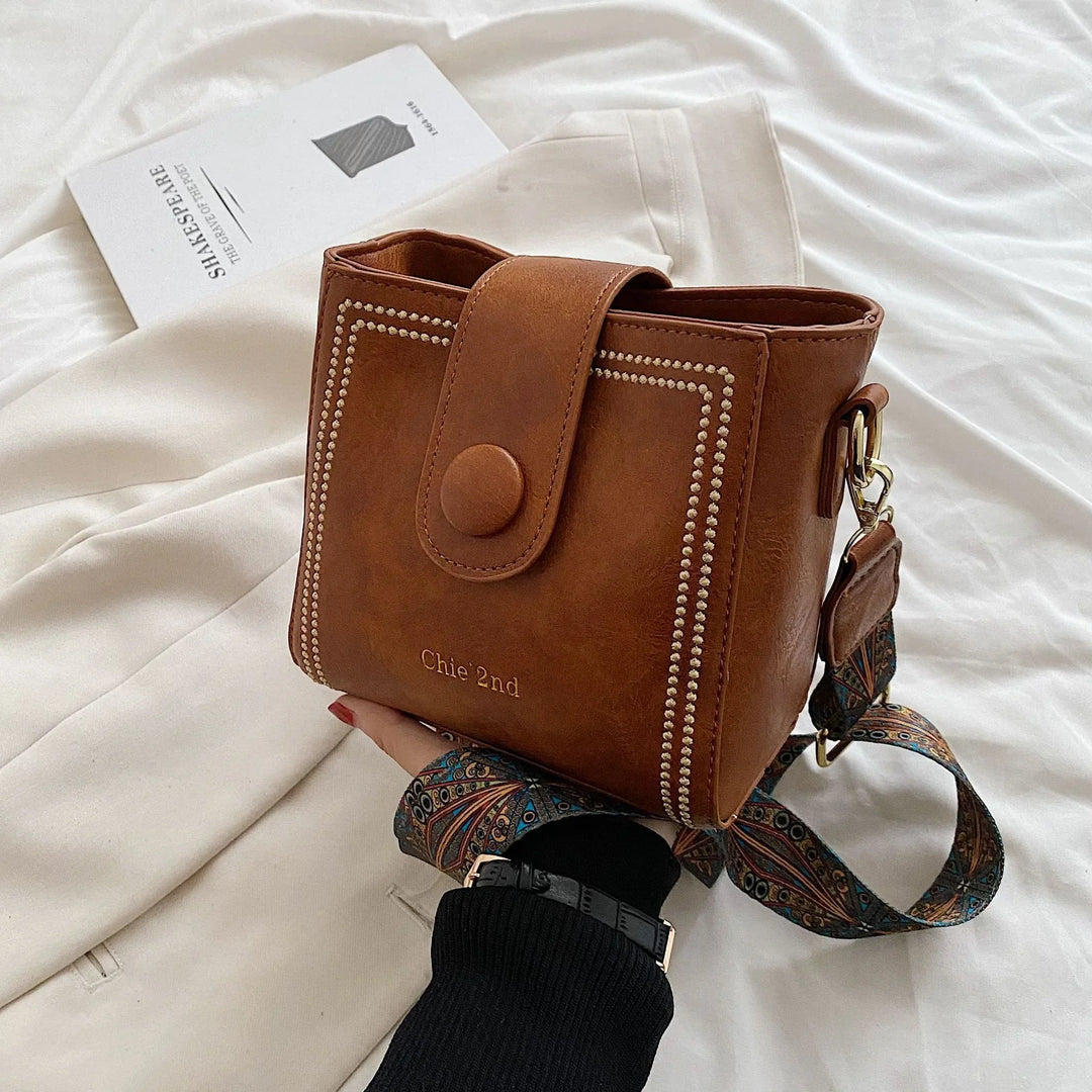 Brown leather crossbody bag with button flap and patterned strap on white fabric background