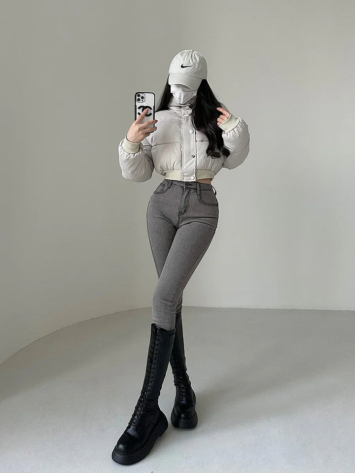 Woman in beige cropped puffer jacket, gray skinny jeans, black knee-high boots, beige cap taking mirror selfie