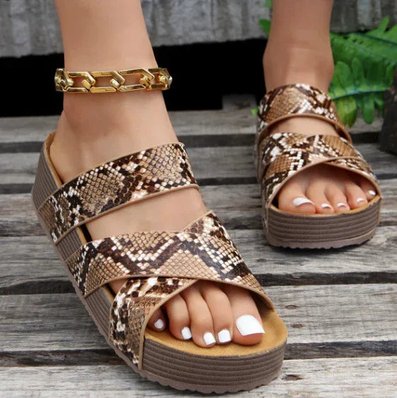 feet wearing snake print wedge sandals with a chunky gold chain anklet on wooden floor