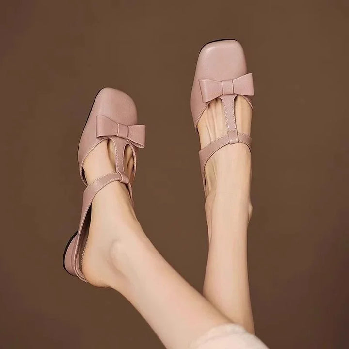 Pink leather T-strap flats with bow detail on feet against brown background