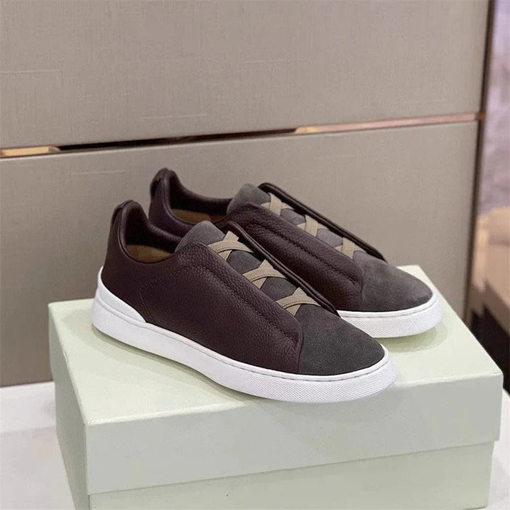 Pair of dark brown and gray leather and suede slip-on sneakers with white soles on a light green box