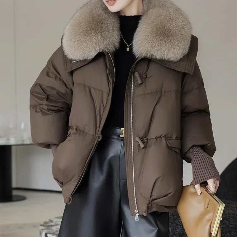 Woman wearing brown puffer jacket with large fur collar and holding tan clutch bag indoors