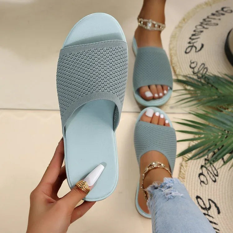 Light blue textured slide sandals worn with white pedicure and gold anklets on tiled floor with decorative mat and plant