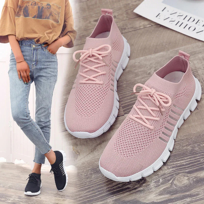 Light pink knitted breathable sneakers with white soles on wooden floor next to model wearing black similar shoes and casual outfit
