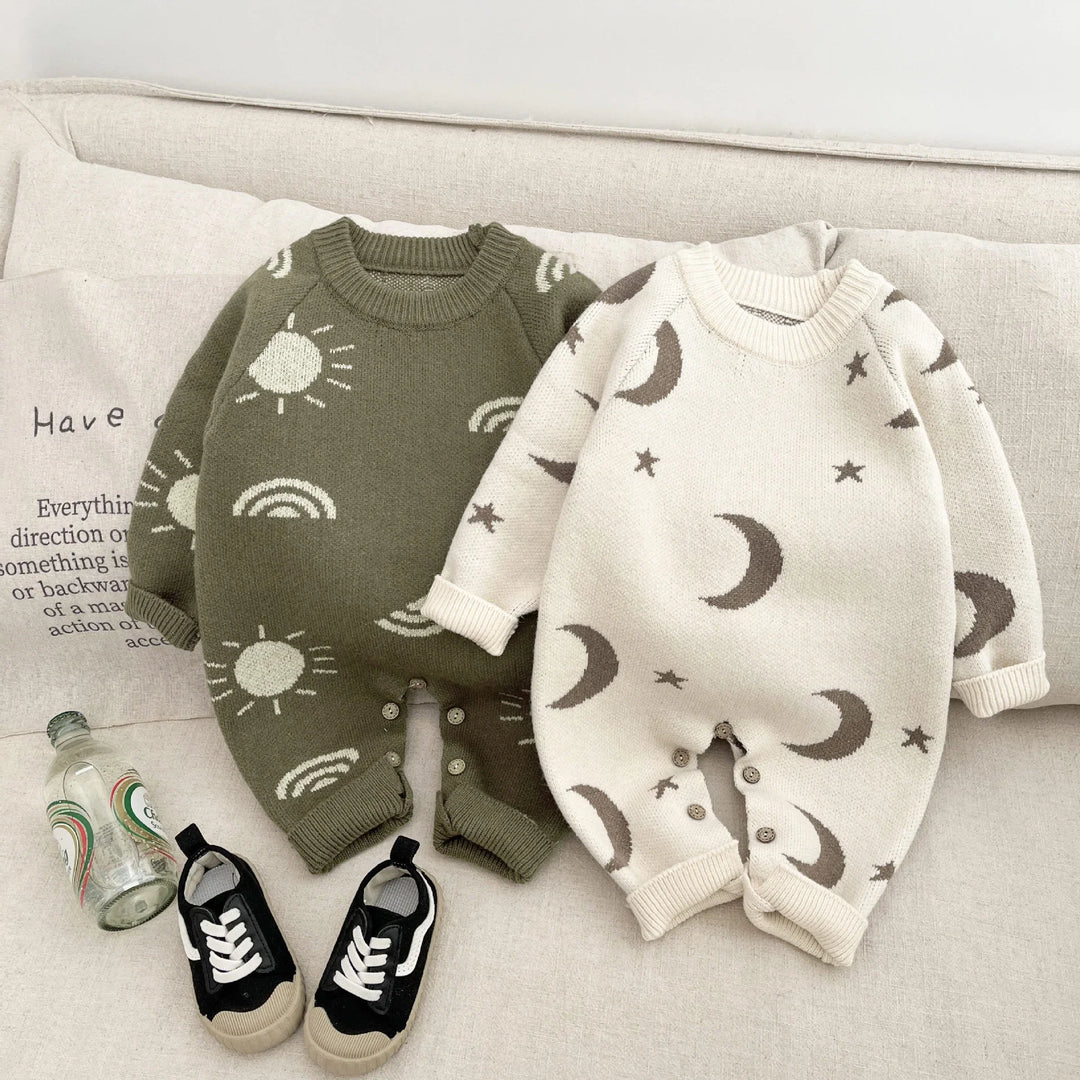 Two knitted baby rompers with moon and sun patterns on a beige couch with black baby sneakers and a glass bottle
