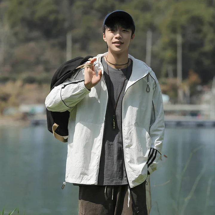 Young man in white jacket and baseball cap carrying backpack outdoors near lake and trees